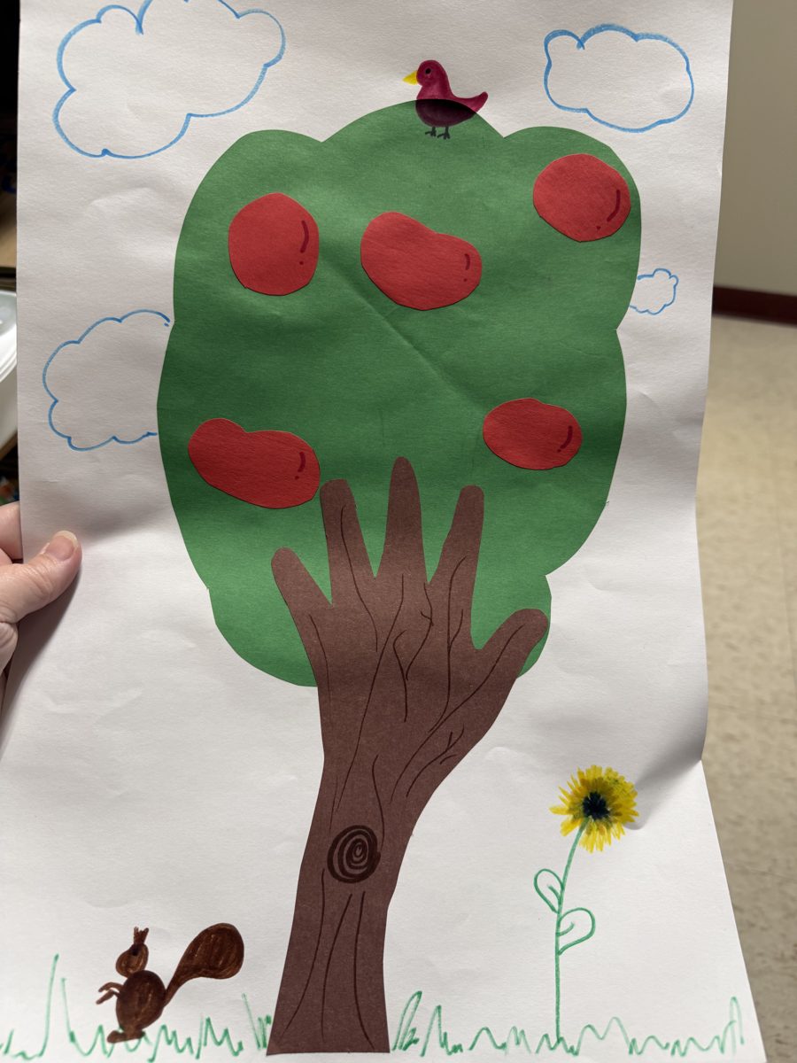 Apple tree craft: a green paper treetop with red apples on a brown handprint trunk, with a bird in the tree, a squirrel, a sunflower and drawn clouds